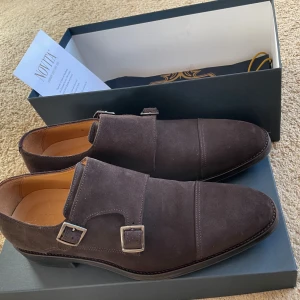 Bruna mocka monkstraps från Novita - Snygga bruna monkstrap-skor från Novita i mjuk mocka med dubbla silverfärgade spännen. Skorna har en elegant spetsig tå och låg klack, perfekt för dig som gillar stilrena och klassiska detaljer. Levereras med originalkartong och dustbag. Köpta för 1700 kr men bara använda under en dag, pga av fel storlek. Se bilderna och bedöm själv.