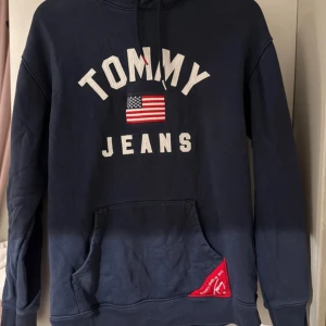 Marinblå hoodie Tommy Jeans M - Snygg marinblå hoodie från Tommy Jeans med broderad logga och amerikansk flagga på bröstet. Tröjan har huva med snörning, stor magficka och en röd detalj med text vid nederkanten. Perfekt för dig som gillar streetwear och klassisk stil.