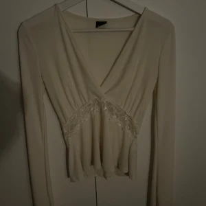 Krämvit omlottblus med spetsdetalj - Säljer en krämvit omlottblus med v-ringning och långa ärmar. Blusen har en fin spetsdetalj under bysten och ett lätt, flödande tyg som ger en feminin siluett. Perfekt för dig som gillar romantiska och eleganta toppar.