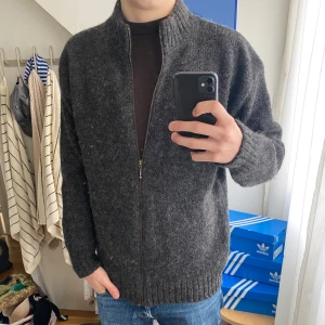 Mörkgrå ullkofta från Ø-Strik - Mysig mörkgrå kofta i ull från Ø-Strik Læsø Uldstue. Koftan har hel dragkedja framtill, ribbade muddar och hög krage. Perfekt för kyliga dagar och enkel att slänga på över en t-shirt eller tröja.