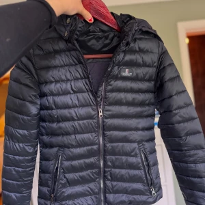 Marin jacka från Kingsland - marin pufferjacka från Kingsland med huva och dragkedja framtill. Jackan har två fickor med dragkedja och quiltad design. Materialet är 100% nylon på utsidan och 100% polyester i fodret. Perfekt för kyliga dagar och snygg streetstil. Jackan är använd max 3/4 gånger och är i jättefint skick. Storlek XS men passar mig som normalt sett har s/m i jackor. Nypris 1899 mitt pris 800.