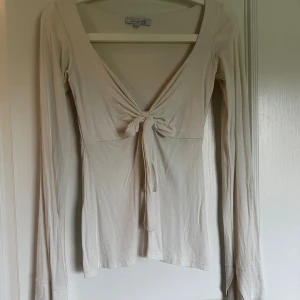 Benvit/vit långärmad topp med knyt - Säljer en vit långärmad topp från Design Si i mjukt bomullsmaterial. Toppen har djup v-ringning och snygg knytning framtill som ger en feminin touch. Passformen är slim och den är perfekt för dig som gillar stilrena och enkla plagg.