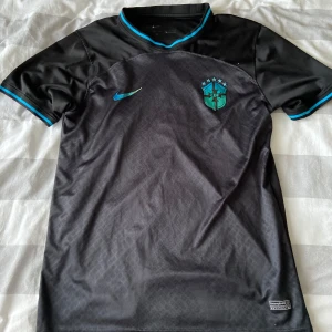 Svart Brasilien Nike fotbollströja - Snygg svart Brasilien fotbollströja från Nike med blå detaljer på krage och ärmslut. Tröjan har korta ärmar, diskret mönster på framsidan och Brasilien-emblem med stjärnor på bröstet. Tillverkad i lätt och ventilerande polyester.