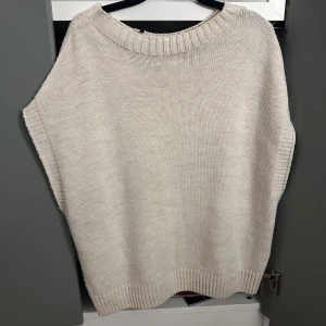 Beige stickad väst med ribbad kant - En stilren beige stickad väst med bred ribbad kant vid halsringning, ärmhål och nederkant. Västen har en rak och loose passform, perfekt att slänga över en t-shirt eller skjorta. Materialet är mjukt och ger en chill vibe till din outfit.