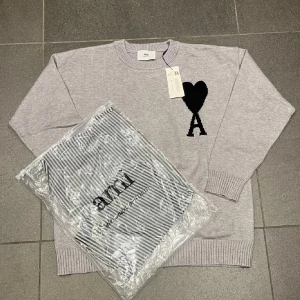 Grå stickad tröja från Ami Paris - Snygg grå stickad tröja från Ami Paris med svart hjärta och A-logga på bröstet. Tröjan har rund hals, ribbade muddar och är långärmad. Perfekt för dig som gillar stilrena och trendiga plagg med en twist.