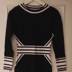 Svartvit randig klänning från H&M - Snygg svart klänning från H&M med vita ränder vid midjan, ärmsluten och halsringningen. Modellen är kroppsnära med lång ärm och kort längd. Perfekt för dig som gillar en sportig och stilren look.
