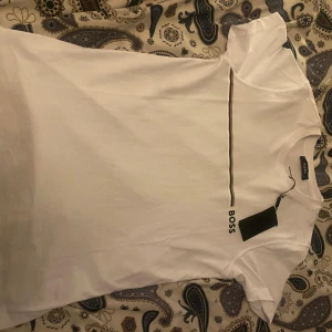 Vit BOSS t-shirt med svart detalj - Vit t-shirt från BOSS i storlek M med en stilren svart linje över bröstet och BOSS-logga tryckt på framsidan. Klassisk rund hals och korta ärmar. Mjuk och skön bomullskvalitet som känns fräsch mot huden. Perfekt för dig som gillar enkel och snygg design.