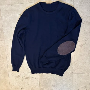 Marinblå stickad tröja Stenströms  - Snygg marinblå stickad tröja från Stenströms med rund hals och ribbade muddar. Tröjan har lång ärm och coola bruna armbågslappar som ger en schysst touch. Perfekt för dig som gillar klassisk stil med lite edge.