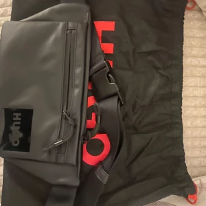 Svart axelväska från Hugo - Snygg svart axelväska från Hugo med stilren design och justerbart band. Väskan har en praktisk dragkedja framtill och en diskret logga i svart på framsidan. Tillverkad i ett tåligt syntetmaterial som ger en modern känsla.