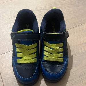 Blå Heelys sneakers med gula detaljer - Säljer ett par coola blå Heelys sneakers med neon-gula snören och detaljer. Skorna har kardborreband och rund tå, samt en platt sula. Perfekta för dig som gillar att rulla fram med stil! Märket syns tydligt på sidan och bak på skon.