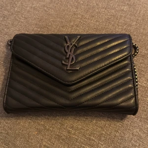 Svart quiltaYSL axelväska i skinn - Snygg svart axelväska från YSL i quiltat skinn med silverfärgad kedja och ikonisk YSL-logga framtill. Väskan har kuvertformad front, magnetlås och ett fack med dragkedja inuti. Perfekt storlek för det viktigaste och ger en lyxig vibe till din outfit.