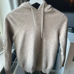 Beige hoodie i ull med huva - Mysig beige hoodie i mjuk ull med huva och snörning. Tröjan har lång ärm och en enkel, stilren design som passar perfekt för kyliga dagar. Ribbstickad kant vid huvan ger en extra touch. Perfekt för dig som gillar en avslappnad och trendig look.