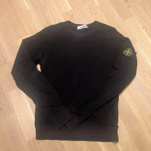 Svart sweatshirt från Stone Island - Svart sweatshirt från Stone Island med klassisk rund hals och ikonisk logotyp-patch på vänster ärm. Tröjan har långa ärmar och ribbade muddar vid ärmslut och nederkant. Perfekt för dig som gillar streetwear och stilren design.