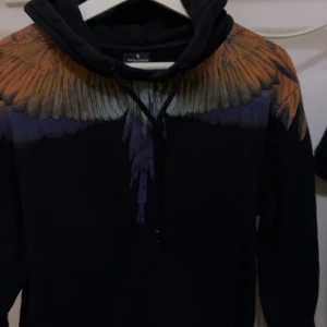 Marcelo Burlon hoodie med fjädertryck - Säljer en svart hoodie från Marcelo Burlon med ikoniskt fjädermönster i orange och lila över axlar och bröst. Tröjan har huva med snörning och är långärmad. Perfekt statement piece för dig som vill sticka ut.