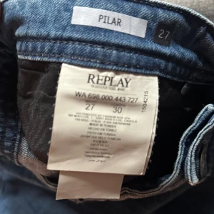 Blå jeansbyxor från Replay, modell Pilar - Snygga blå jeansbyxor från Replay i modellen Pilar. Jeansen har klassisk femficksdesign, knäppning med fyra knappar framtill och coola detaljer vid midjan. Materialet är mjukt jeans och passformen är normal med raka ben. Perfekta för dig som gillar stilrena och bekväma jeans.
