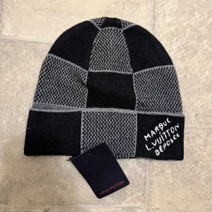 Rutig mössa från Louis Vuitton - Snygg rutig mössa från Louis Vuitton i svart och grått. Mössan är stickad i en lyxig mix av kashmir, ull och silke. Den har ett coolt rutmönster och broderad text framtill. Perfekt accessoar för att lyfta din vinterstil.