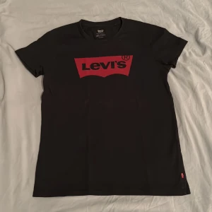 Svart Levi's t-shirt med rött tryck XS - Svart t-shirt från Levi's i storlek XS med klassiskt rött logotryck på bröstet. T-shirten har rund halsringning och korta ärmar. Tillverkad i mjuk bomull för en skön känsla. Perfekt för en avslappnad och stilren look.