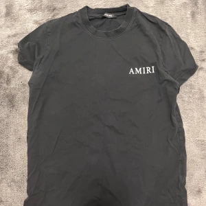 Svart AMIRI t-shirt med ängeltryck - Svart t-shirt från AMIRI med vit logga på bröstet och stort tryck av en ängel på ryggen. Klassisk rund hals och korta ärmar. Snygg och stilren design som sticker ut med det detaljerade motivet på baksidan. ( som du ser i bilden så finns det små vita fläckar , det är endast i bilden inte om du ser den framför dig.  Passar perfekt för dig som är 15 år och väger cirka 60-70 kg.
