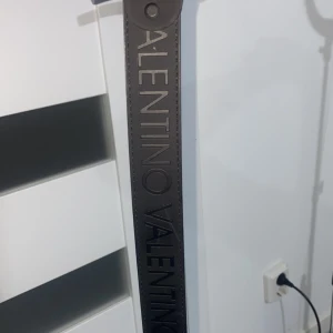 Svart Valentino bälte - Snyggt svart bälte från Valentino med präglad logotyp längs hela remmen. Bältet är tillverkat i skinn och har en stilren, rak form som passar till jeans eller kostymbyxor. Perfekt accessoar för att lyfta din outfit med en lyxig touch. (Pris kan diskuteras)