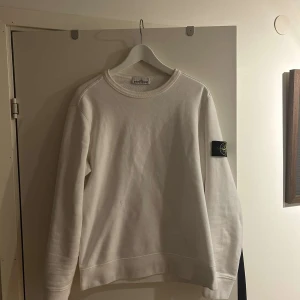 Vit sweatshirt från Stone Island - Vit sweatshirt från Stone Island med klassisk rund hals och lång ärm. Tröjan har den ikoniska svarta patchen med logga på vänster ärm. Mjuk bomullskvalitet och enkel, clean design som passar perfekt till jeans eller cargo.