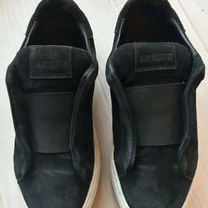 Svarta sneakers från Axel Arigato - Snygga svarta sneakers från Axel Arigato med stilren design och bred elastisk rem över foten. Ovandel i mjuk mocka och vit platt gummisula. Minimalistisk look utan snörning, perfekt för dig som gillar cleana och moderna skor.