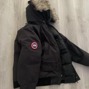 Svart dunjacka från Canada Goose - Säljer en svart dunjacka från Canada Goose med pälskantad huva och klassisk logga på ärmen. Jackan har stora fickor framtill, dragkedja och ribbade muddar. Perfekt för kalla vinterdagar och riktigt snygg streetstil.
