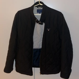 Svart quiltad jacka från GANT - Quiltad svart jacka från GANT med ståkrage och dragkedja framtill. Jackan har ett diskret GANT-emblem på bröstet och snyggt rutmönster. Insidan är randig i blått och vitt, vilket ger en fräsch kontrast. Perfekt för dig som gillar stilren och klassisk look.