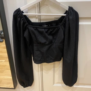 Svart croppad blus från Bik Bok - Säljer en svart croppad blus från Bik Bok med puffiga långärmar och fyrkantig urringning. Toppen har en lätt textur i tyget och elastiska detaljer vid axlar och ärmslut för en snygg passform. Perfekt att styla med höga jeans eller kjol.