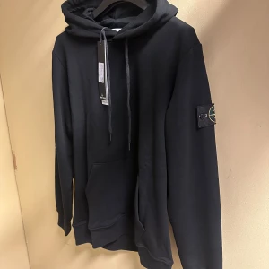 Svart hoodie från Stone Island - Svart hoodie från Stone Island med klassisk patch på vänster ärm. Tröjan har huva med snörning, stor magficka och ribbade muddar. Perfekt för dig som gillar streetwear och vill ha en stilren look. Materialet är mjuk bomull och passformen är normal.