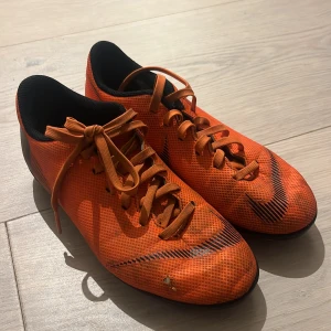 Nike Mercurial orange fotbollsskor - Säljer ett par Nike Mercurial fotbollsskor i en riktigt snygg orange färg med svarta detaljer och snörning. Skorna har en låg profil och är tillverkade i syntetmaterial för lätt känsla på planen. Perfekt för dig som vill sticka ut på matchen!