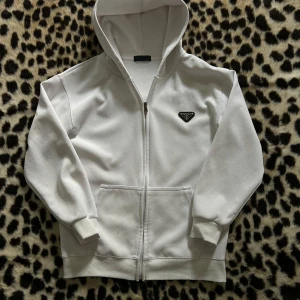 Prada Hoodie - Snygg vit hoodie från Prada med dragkedja framtill och klassisk huva. Tröjan har två fickor på magen och en svart Prada-logga på bröstet. Perfekt för en clean och stilren look. Tröjan är i mycket bra skick. Priset går att diskutera😇