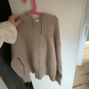 Beige brun  stickad hoodie med omlottkrage - Mysig beige stickad hoodie med omlottkrage och snörning i halsen. Tröjan har långa ärmar och en relaxed fit, perfekt för kyliga dagar. Enkel och stilren design som passar till många outfits.