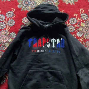 Svart Trapstar hoodie med tryck - Svart hoodie från Trapstar med stor huva och fett tryck i blått, rött och vitt på bröstet. Texten 'IT'S A SECRET' under loggan. Mjuk och skön, perfekt för chill eller streetwear. Klassisk känguruficka framtill och dragsko i huvan.