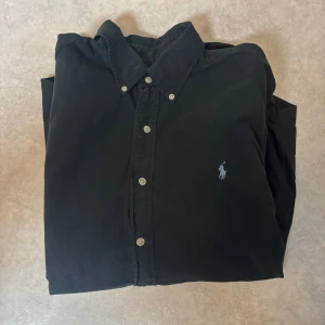 Svart skjorta från Ralph Lauren L - Svart skjorta från Ralph Lauren i slim fit med klassisk krage och knappar framtill. Skjortan har lång ärm och den ikoniska broderade Polo-loggan på bröstet. Tillverkad i mjuk bomull som känns skön mot huden. Perfekt för dig som gillar stilrena och tidlösa plagg.