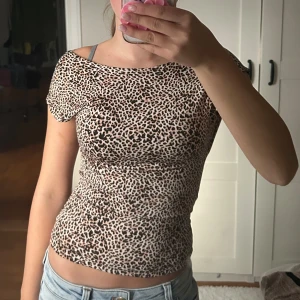 Leopardmönstrad topp med öppen rygg - Säljer en slim leopardmönstrad topp med korta ärmar och öppen rygg. Toppen har en beige bas med svarta och bruna leopardfläckar, är figurnära och har en snygg offshoulder-look. Perfekt för dig som gillar djurmönster och vill sticka ut.