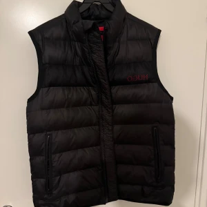 Svart dunväst från Hugo Boss - Svart dunväst från Hugo Boss med quiltad design och hög krage. Västen har två praktiska fickor med dragkedja och ett diskret rött Hugo Boss-tryck på bröstet. Materialet är syntet och polyester, vilket gör den lätt och bekväm. Perfekt för lager-på-lager under kyliga dagar. Litet skavanker på sista bilden därför lägre pris 
