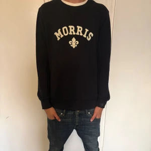 Svart sweatshirt från Morris, storlek M - Svart sweatshirt från Morris med vit broderad logga och emblem på bröstet. Tröjan är i mjuk bomull och har en avslappnad passform som funkar till jeans eller shorts. Hör av er vid minsta fundering!