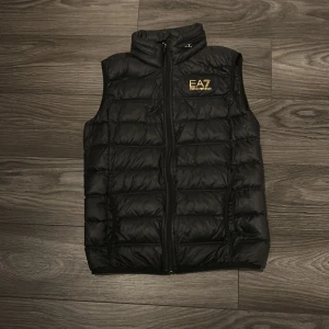Svart dunväst från EA7 Emporio Armani - Snygg svart dunväst från EA7 Emporio Armani med guldfärgat logotryck på bröstet. Västen har quiltad design, hög krage och dragkedja framtill. Perfekt för lager-på-lager och streetstyle. Passar dig som gillar exklusiva märken och stilren look.