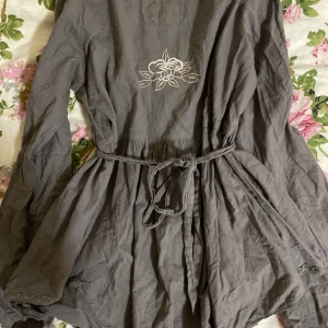 Grå kortklänning från Tina Wodstrup - Supersöt grå kortklänning från Tina Wodstrup med långa ärmar, knappar framtill och broderad blomdetalj på ryggen. Klänningen har knytband i midjan, spetsdetaljer vid ärmslut och nederkant samt veckad överdel för extra stil.