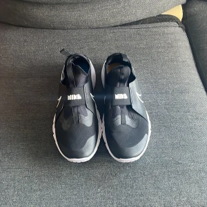 Svarta Nike sneakers med vit sula - Säljer ett par svarta Nike sportskor med bred vit sula och ett 'Just Do It'-tryck på sidan. Skorna har elastiskt band över foten med Nike-logga och är tillverkade i syntetmaterial med meshdetaljer för extra komfort. 