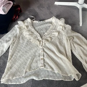 Vit blus med volang från Zara - Supersöt vit blus från Zara med prickigt mönster, volangdetaljer längs v-ringningen och knappar framtill. Långärmad med smock vid ärmslut och transparent, lätt material som ger en luftig känsla. Perfekt för dig som gillar romantisk och feminin stil.
