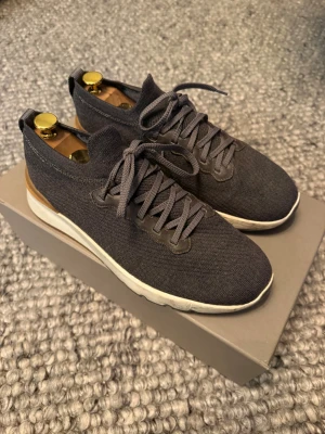Brunello Cucinelli Sneakers - Säljer ett par stilrena grå sneakers från Brunello Cucinelli med stickad ovandel, snörning och vita samt bruna detaljer på sulan. Skorna har en sportig vibe och är perfekta för dig som gillar exklusiva och bekväma sneakers med modern design.