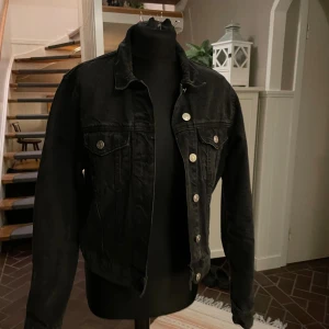 Svart jeansjacka med knappar - Snygg svart jeansjacka med silverfärgade knappar framtill. Jackan har två bröstfickor med lock och knapp, samt justerbara knappar i midjan. Perfekt för dig som gillar en cool och enkel stil.