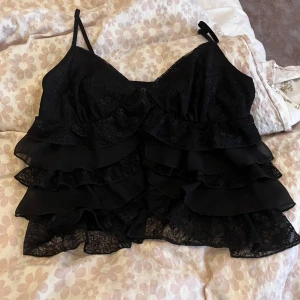 Svart volangig spets-linnetopp - Säljer en svart linne-topp med tunna axelband och flera lager volanger i spets och tyg. Toppen har en feminin och cool look med detaljerad spets och volanger som ger extra attityd. Perfekt för dig som gillar att sticka ut med din stil.