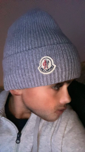 Grå ribbstickad mössa från Moncler - Snygg grå ribbstickad mössa från Moncler med klassisk Moncler-logga framtill. Mössan har uppvikt kant och är tillverkad i mjukt ullmaterial som håller dig varm. Perfekt för dig som vill ha en clean och stilren look med exklusiv känsla.