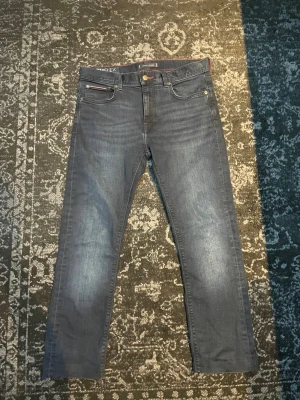 Mörkblå slim jeans från Tommy Hilfiger - Snygga mörkblå jeans från Tommy Hilfiger med diskreta slitningar och röda sömdetaljer. Klassisk femficksmodell med logotyp på bakfickan. Stretchigt material för skön passform och raka ben.
