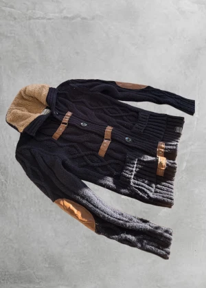 Mörkblå fur cardigan jacka från Tazzio - Mörkblå stickad fur cardigan från Tazzio med grov kabelstickning, bruna detaljer i skinn på armbågar och knäppning, samt mjuk beige teddyfleece i kragen. Jackan har stora fickor och både knappar och spännen framtill. Perfekt för kalla dagar.