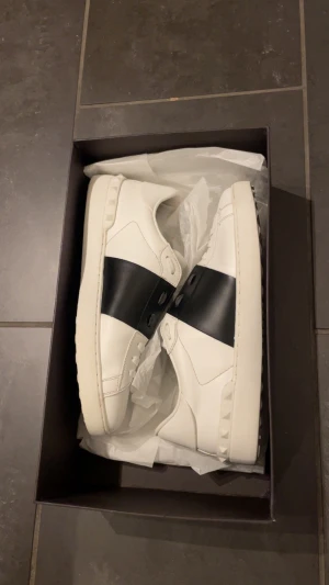 Valentino Garavani Open sneakers vit/svart - Säljer ett par Valentino Garavani Open sneakers i vitt skinn med bred svart läderdetalj över mitten. Snygga nitar längs sidorna och platt sula med mönstrad undersida. Klassisk rund tå och snörning framtill. Perfekt för dig som gillar stilrena designer. Självfallet äkta, Kan bevisa i chatten 