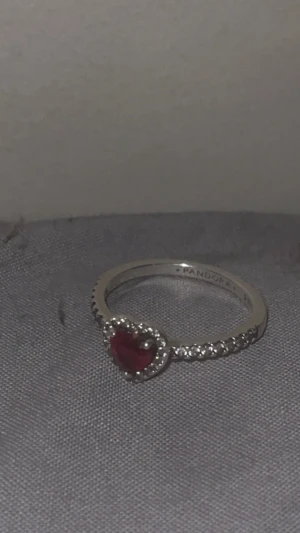 Pandora silverring med röd hjärtsten - Superfin silverring från Pandora med en röd hjärtformad sten i mitten, omgiven av små glittrande stenar längs ringen. Perfekt för dig som gillar romantiska detaljer och vill ha något som sticker ut.
