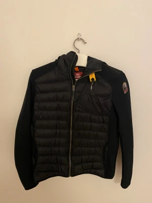 Svart pufferjacka Parajumpers  - Svart pufferjacka från Parajumpers med quiltad front och ärmar i stickad struktur. Jackan har dragkedja, huva och den klassiska PJS-loggan på ärmen. Snygga detaljer med gul rem vid halsen och orange sömmar. Perfekt för kyliga kvällar.